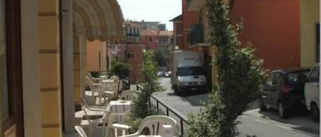 Una calle estrecha con coches aparcados y un camión, con mesas al aire libre y sillas de plástico blanco, y edificios con balcones.