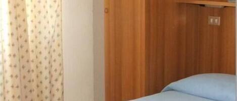 Una cama individual con una colcha azul claro en una habitación con un armario de madera y una cortina de estampado pequeño.