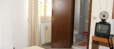 Una habitación con una cama, una puerta de madera, un inodoro y un televisor sobre un mueble de madera.