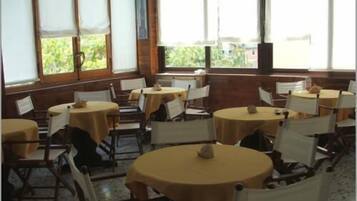Un espace repas avec des tables et des chaises rondes, de grandes fenêtres équipées de stores et une vue sur la verdure environnante.