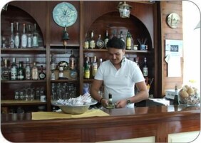 Zona de bar con estantes de madera llenos de botellas y vasos, un reloj y una barra donde una persona prepara comida.