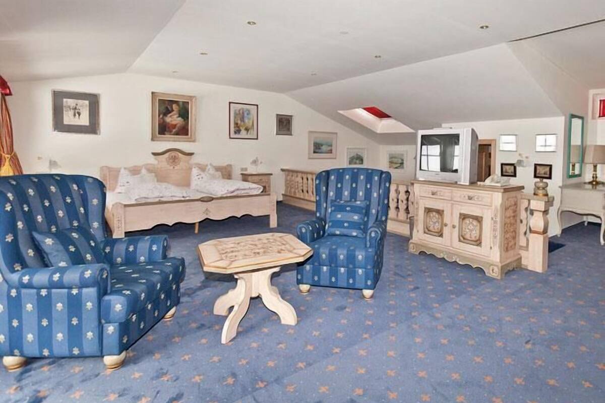 A room with a bed, two blue armchairs, a small table, and a TV on a stand.