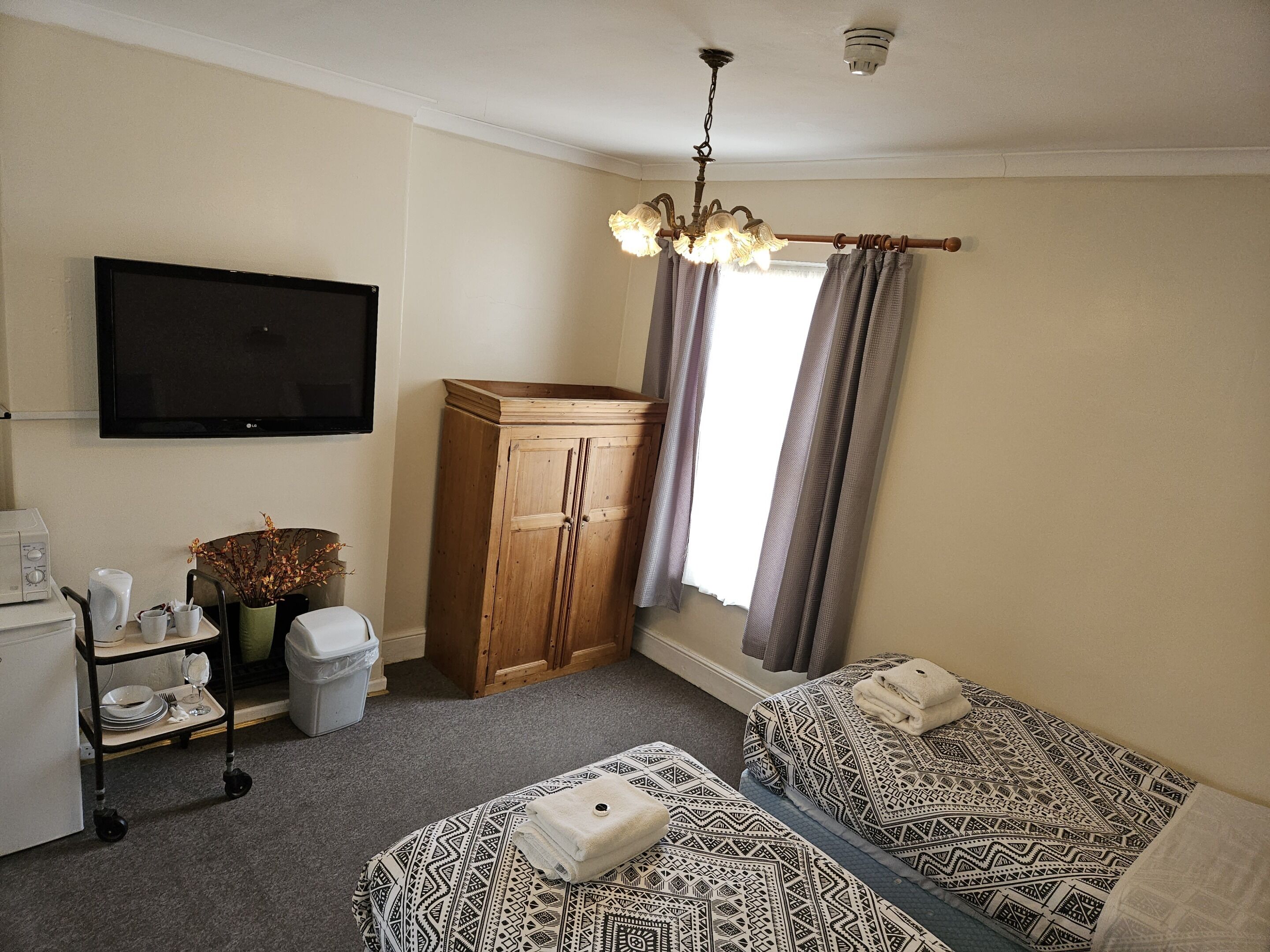 Una habitación con dos camas, televisión, un mueble de madera, una mesita con un jarrón y una ventana con cortinas.