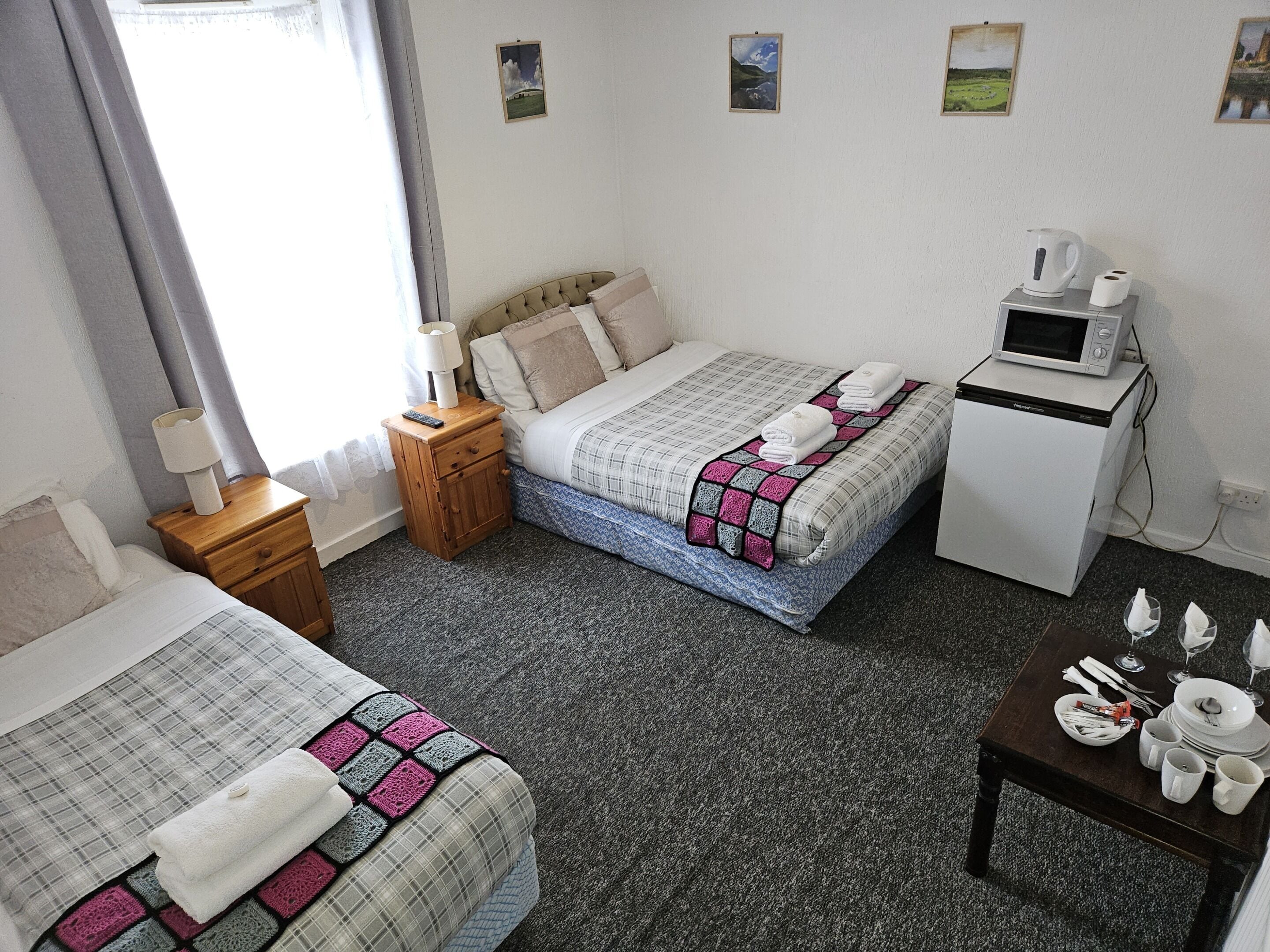A hotel room with two beds, a bedside table with a lamp, a small wooden nightstand, a microwave, and a table set with plates, glasses, and cutlery.