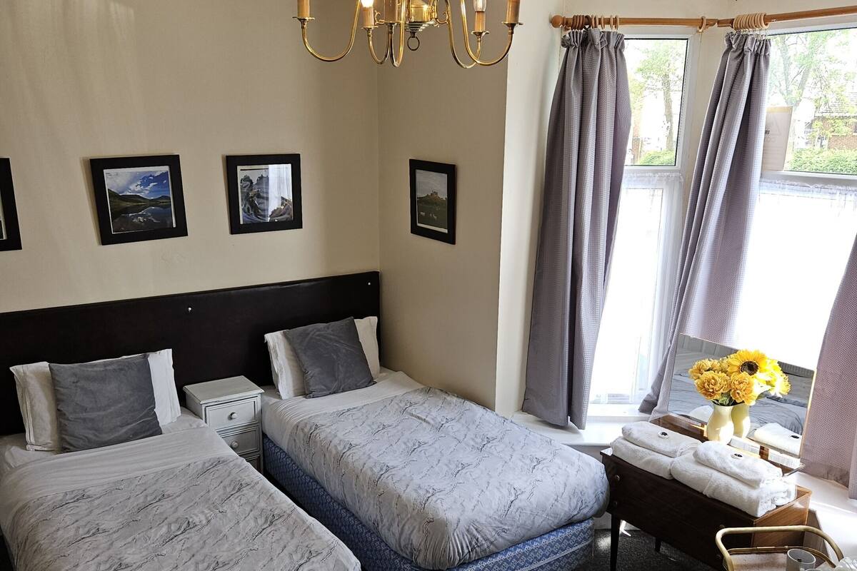 A hotel room with two beds, a chandelier, a bedside table with a vase of flowers, and a window with curtains.
