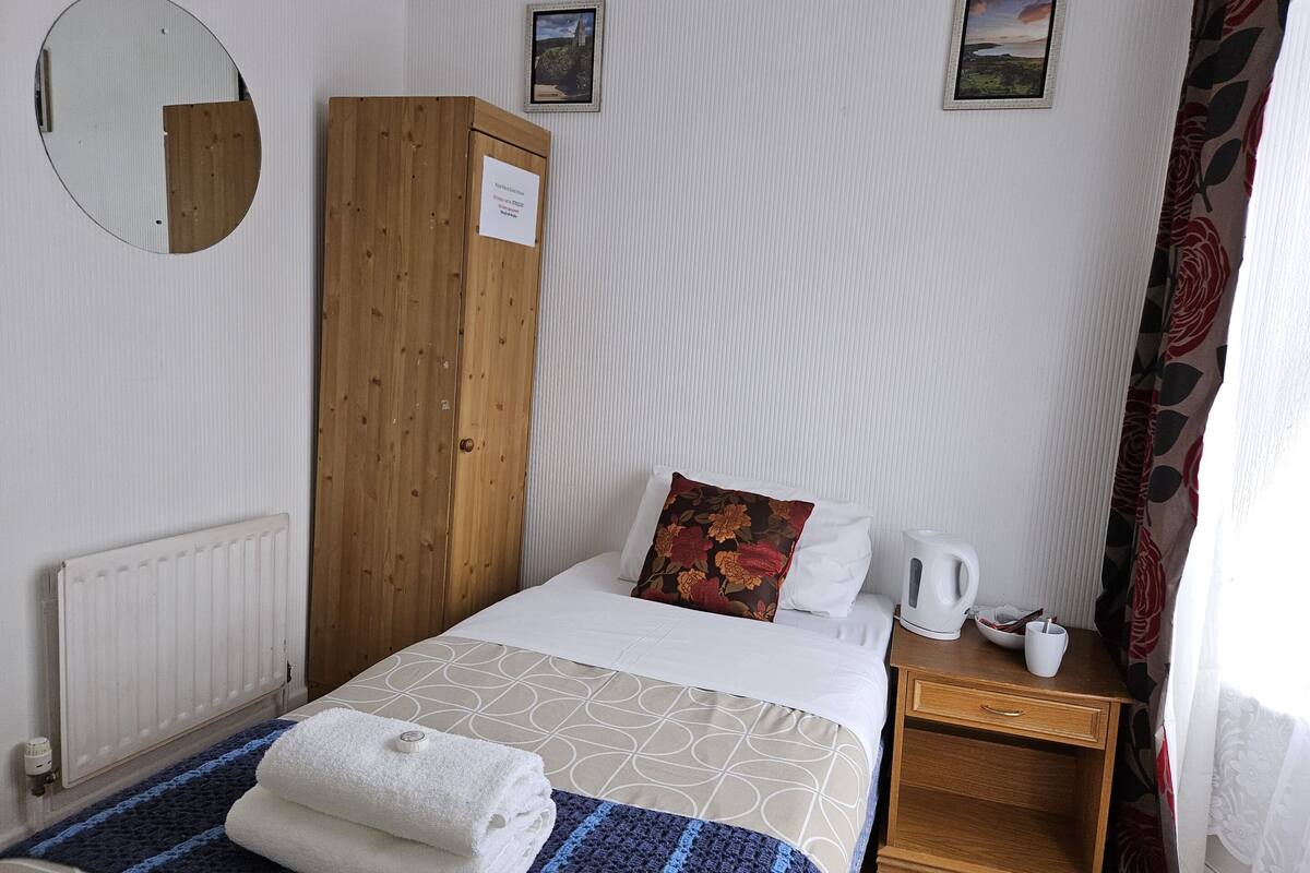 A neatly arranged hotel room with a bed, a wooden wardrobe, a radiator, a small table with a kettle, and two framed pictures on the wall.