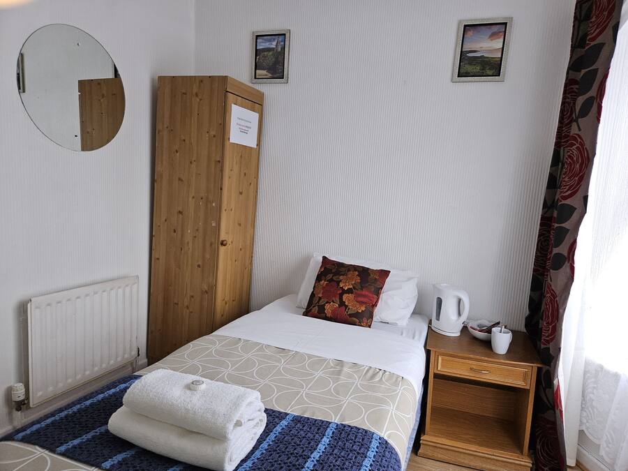 A neatly arranged hotel room with a bed, a wooden wardrobe, a radiator, a small table with a kettle, and two framed pictures on the wall.