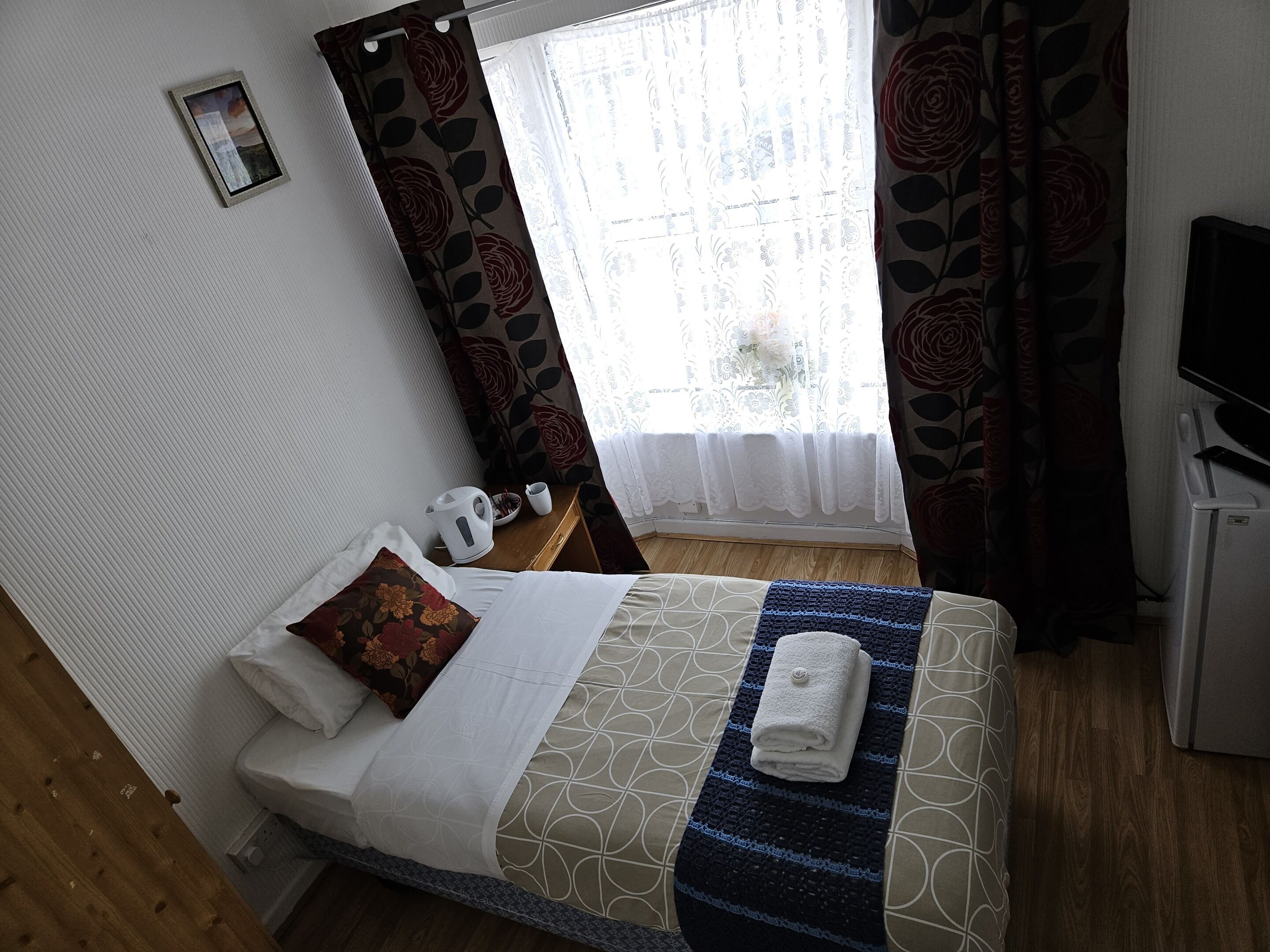 A neatly made bed with a patterned bedspread, a folded towel, and a small bedside table with a kettle and a cup.
