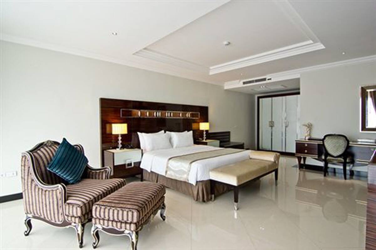 A hotel room with a large bed, a sitting area with a chair and ottoman, a desk with a chair, and a mirror.