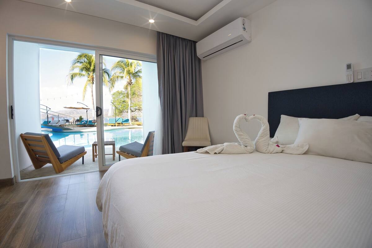 A hotel room with a large bed, a view of a pool, and a balcony with chairs.
