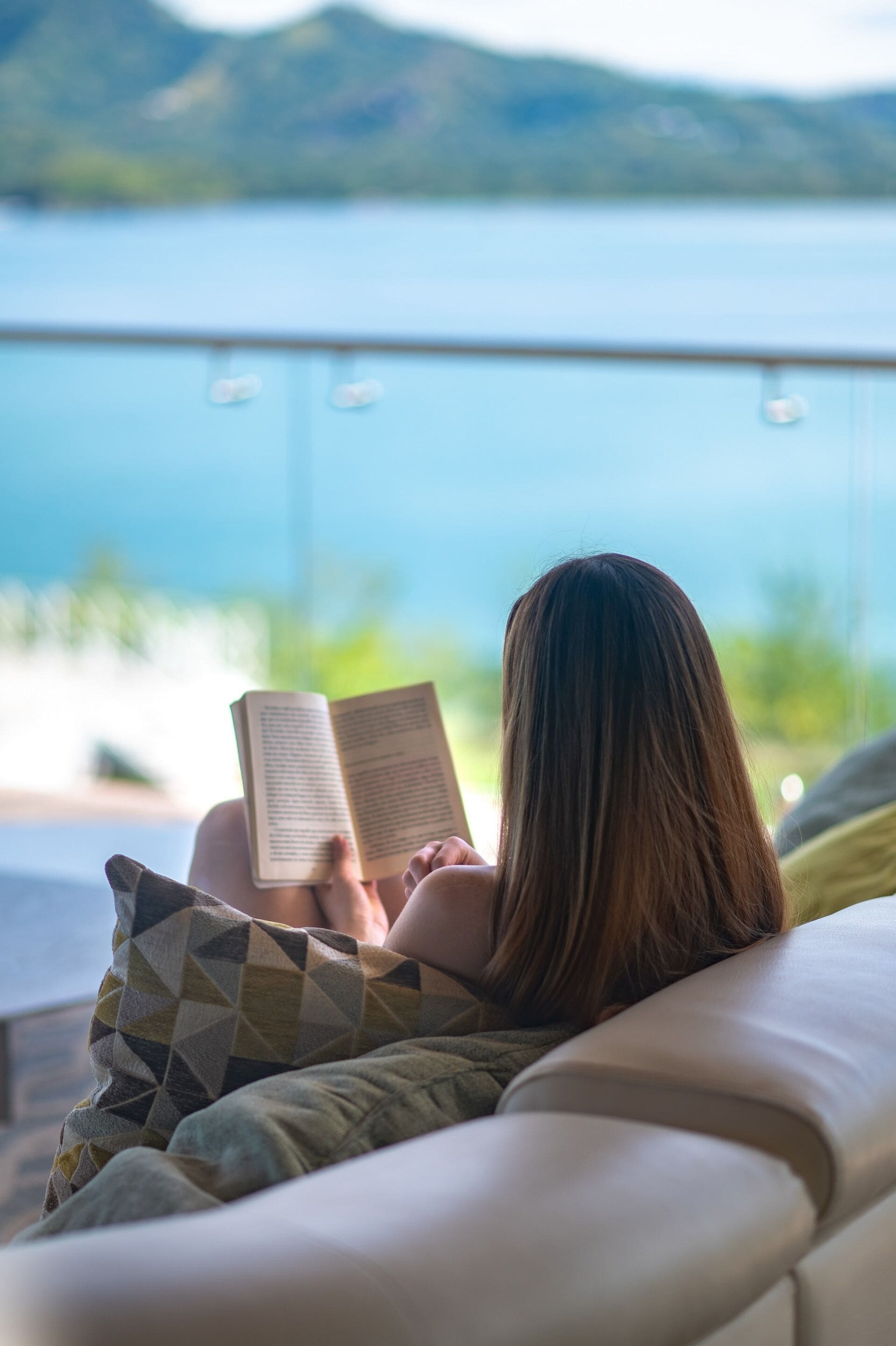 Uma pessoa lendo um livro em um sofá, com vista para um corpo de água e montanhas ao fundo.