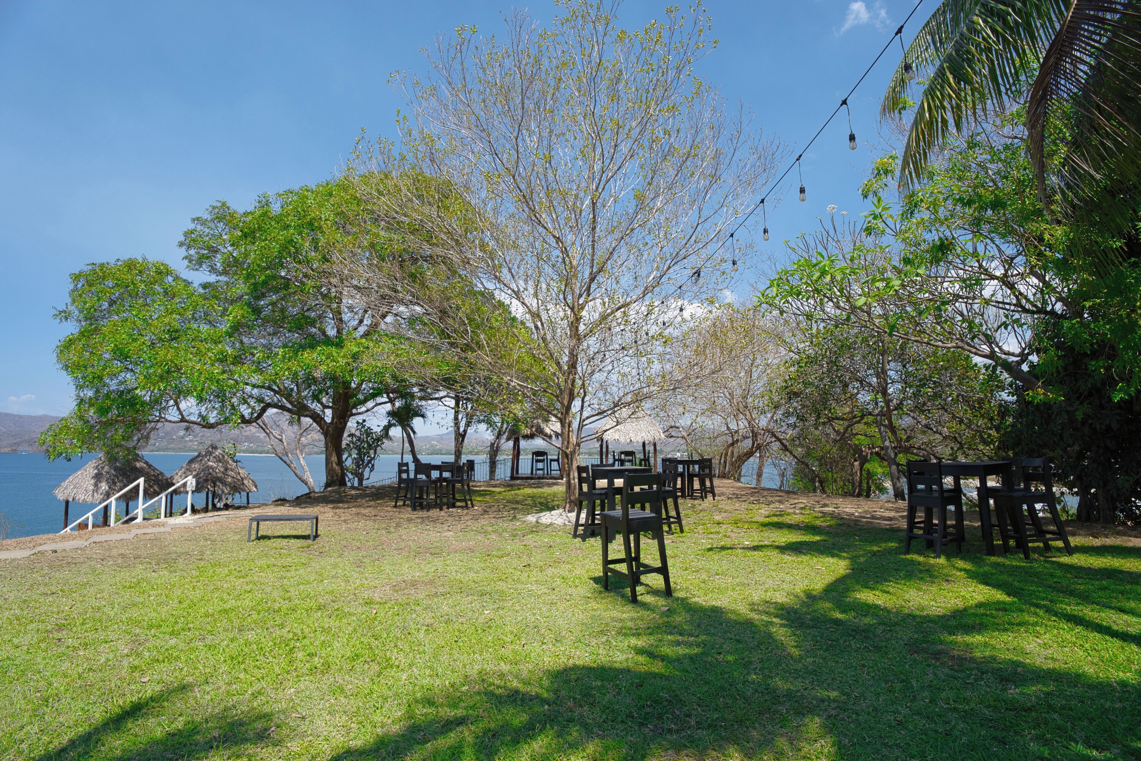 An outdoor seating area with wooden tables and chairs, a thatched-roof structure, and a view of a body of water.