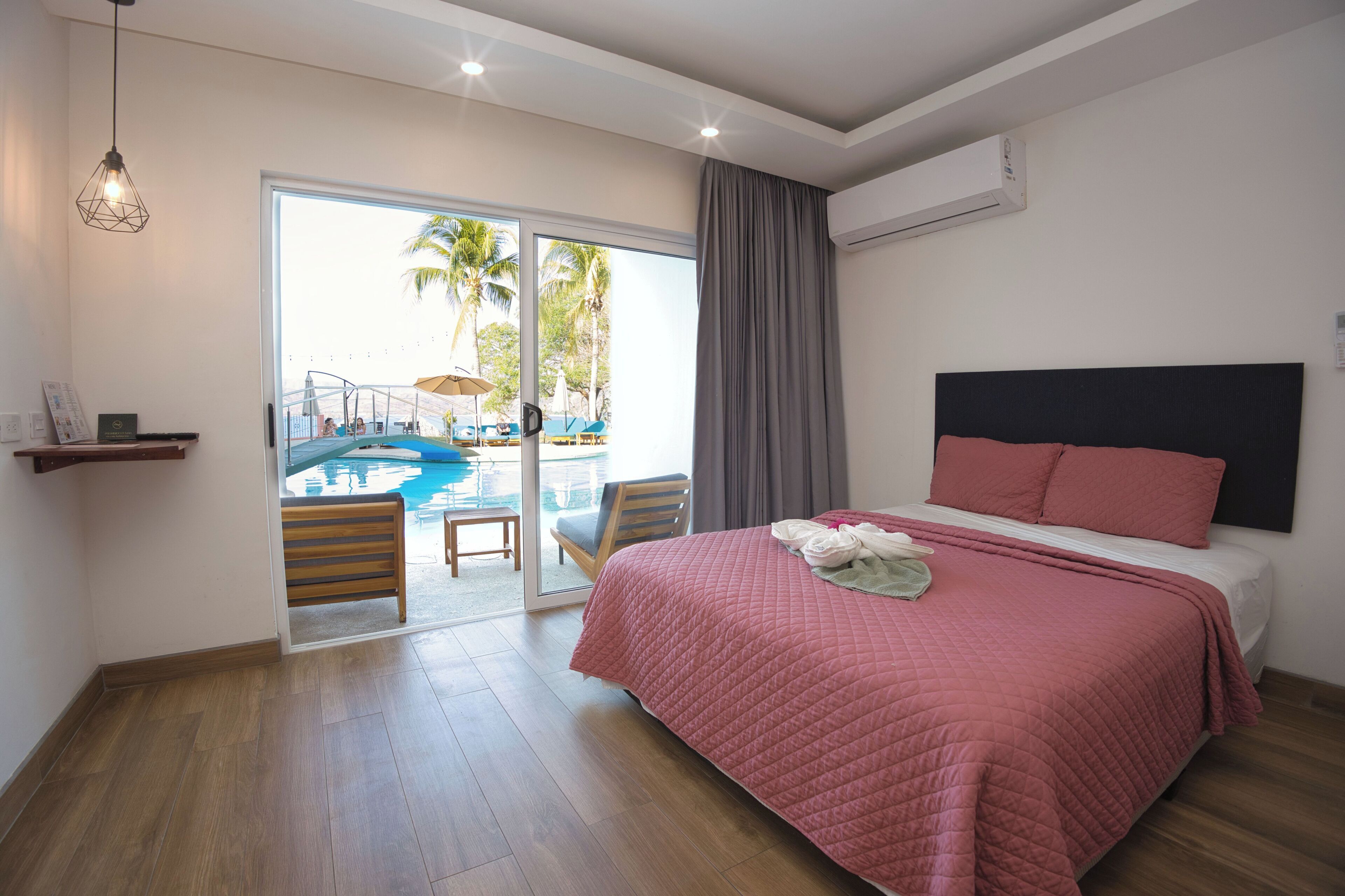 A hotel room with a large bed, a view of a pool, a balcony, and a wooden floor.