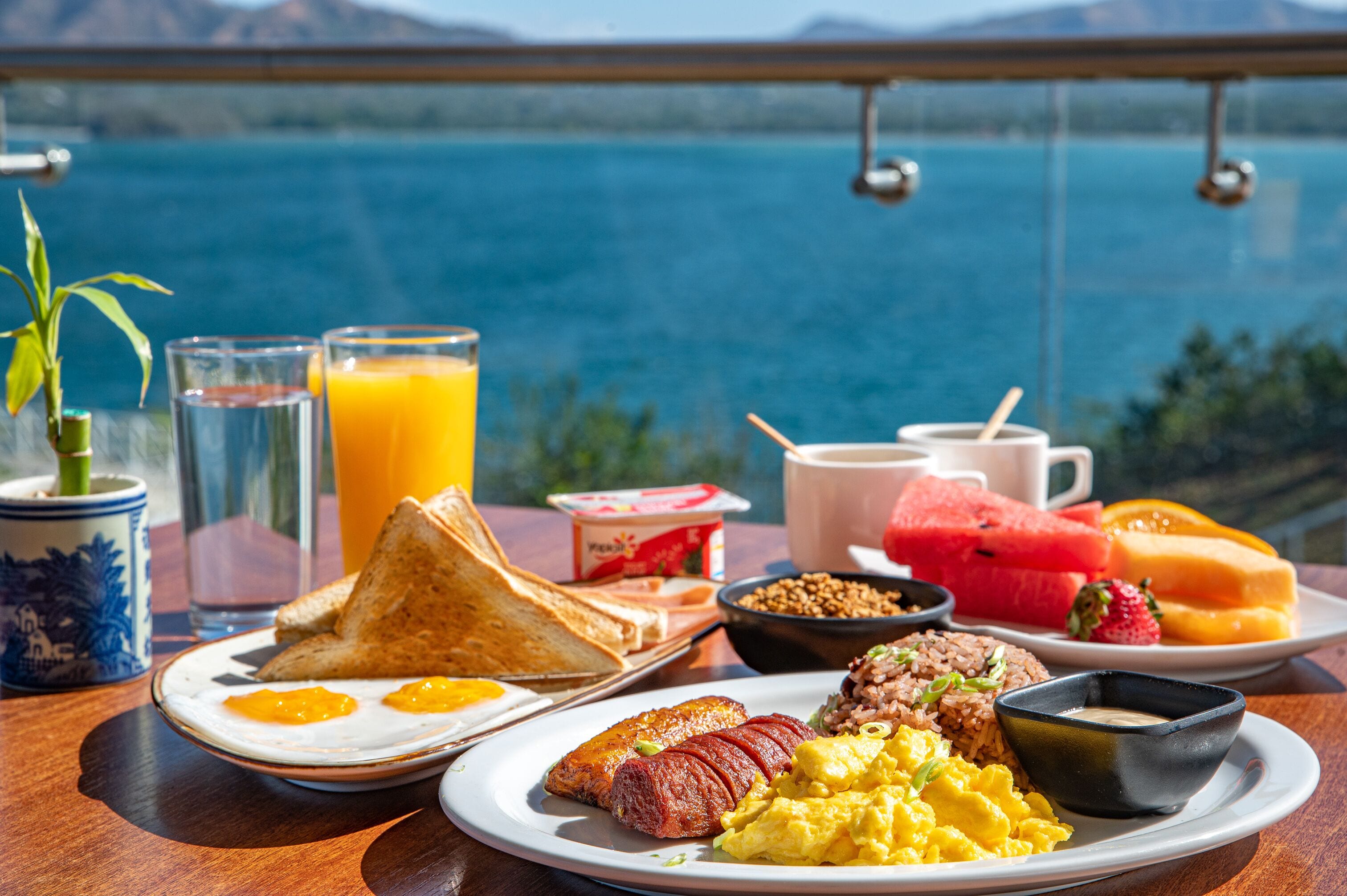 A breakfast table with toast, eggs, sausages, and fruits, with a scenic view of a lake.