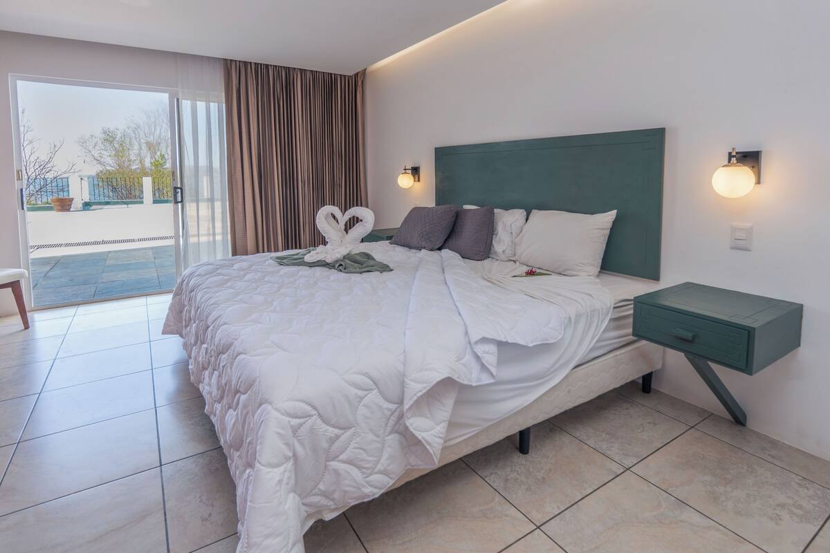 A hotel room with a large bed, a green headboard, two bedside tables with lamps, and a view of the outdoors through a sliding glass door.