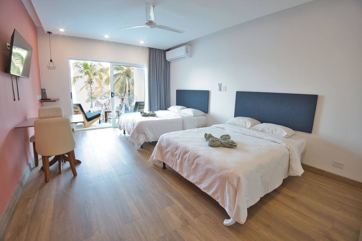 A modern hotel room with two beds, a flat-screen TV, a ceiling fan, and a view of palm trees.