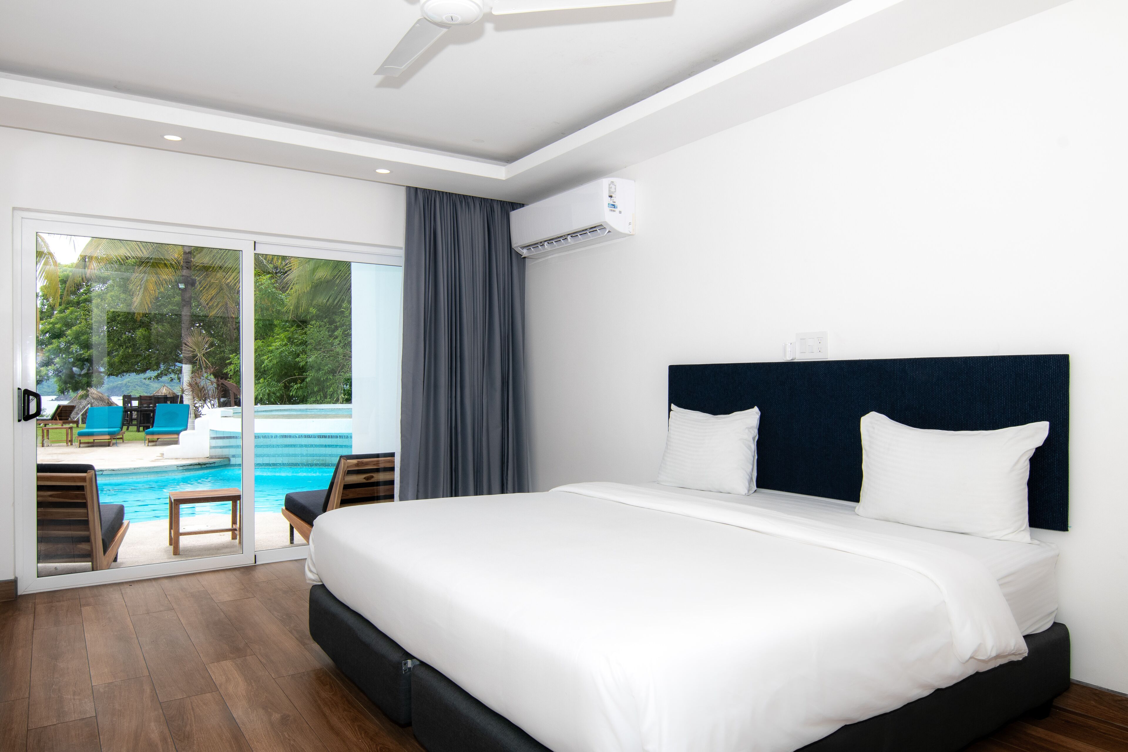 A hotel room with a large bed, a ceiling fan, an air conditioning unit, and a view of a pool and outdoor seating area.