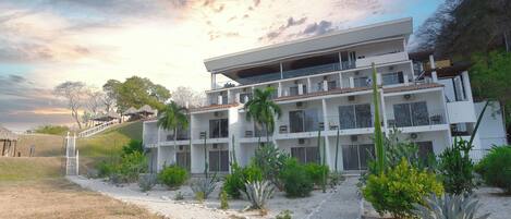 A modern multi-story hotel with a flat roof, balconies, and a landscaped garden with various plants.