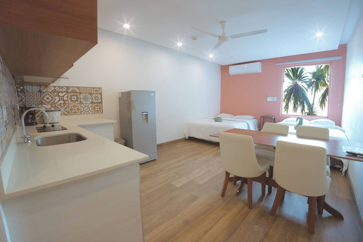A compact hotel room with a kitchenette, a bed, a dining area with chairs, a ceiling fan, and a window with a view of palm trees.