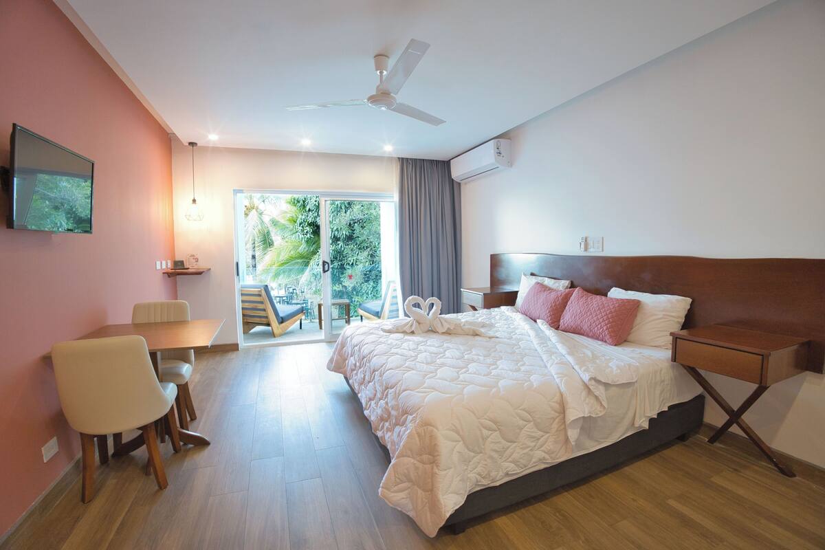 A hotel room with a bed, a desk, a chair, a ceiling fan, and a view of the outdoors.