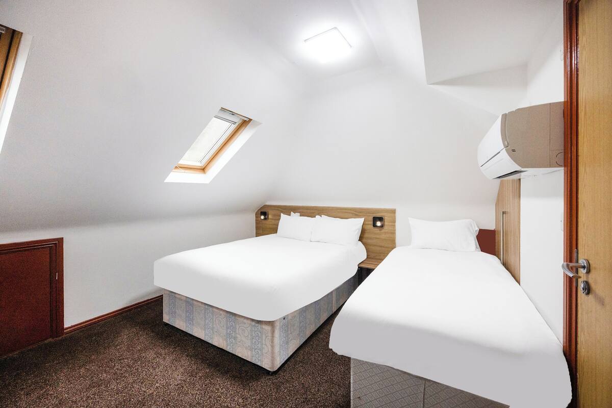 Une chambre d’hôtel avec deux lits, une lucarne, un ventilateur de plafond et une armoire en bois.