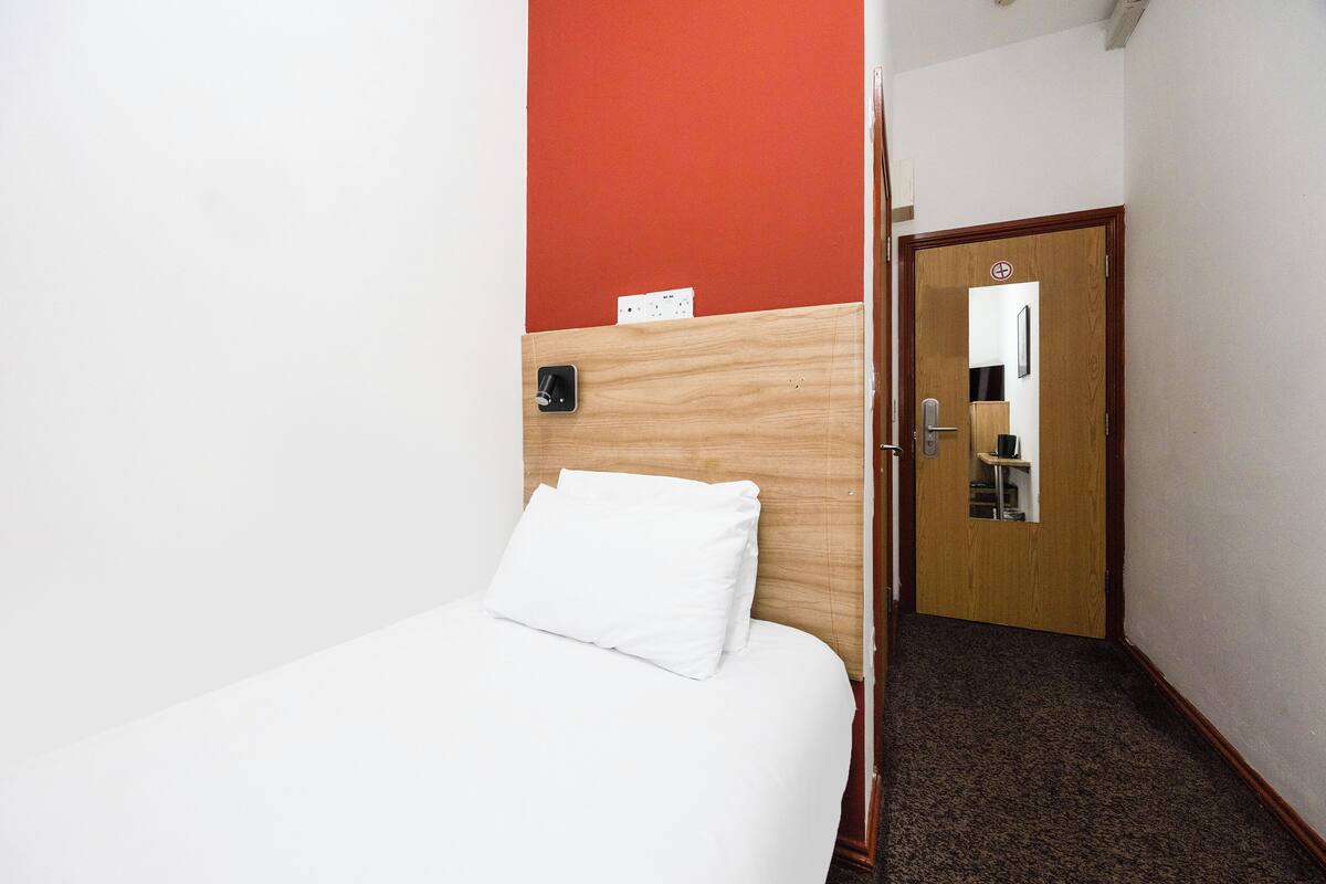 Une chambre d’hôtel avec une tête de lit en bois, du linge de lit blanc, un mur d’accent rouge, et une porte dotée d’un miroir et d’une étagère.