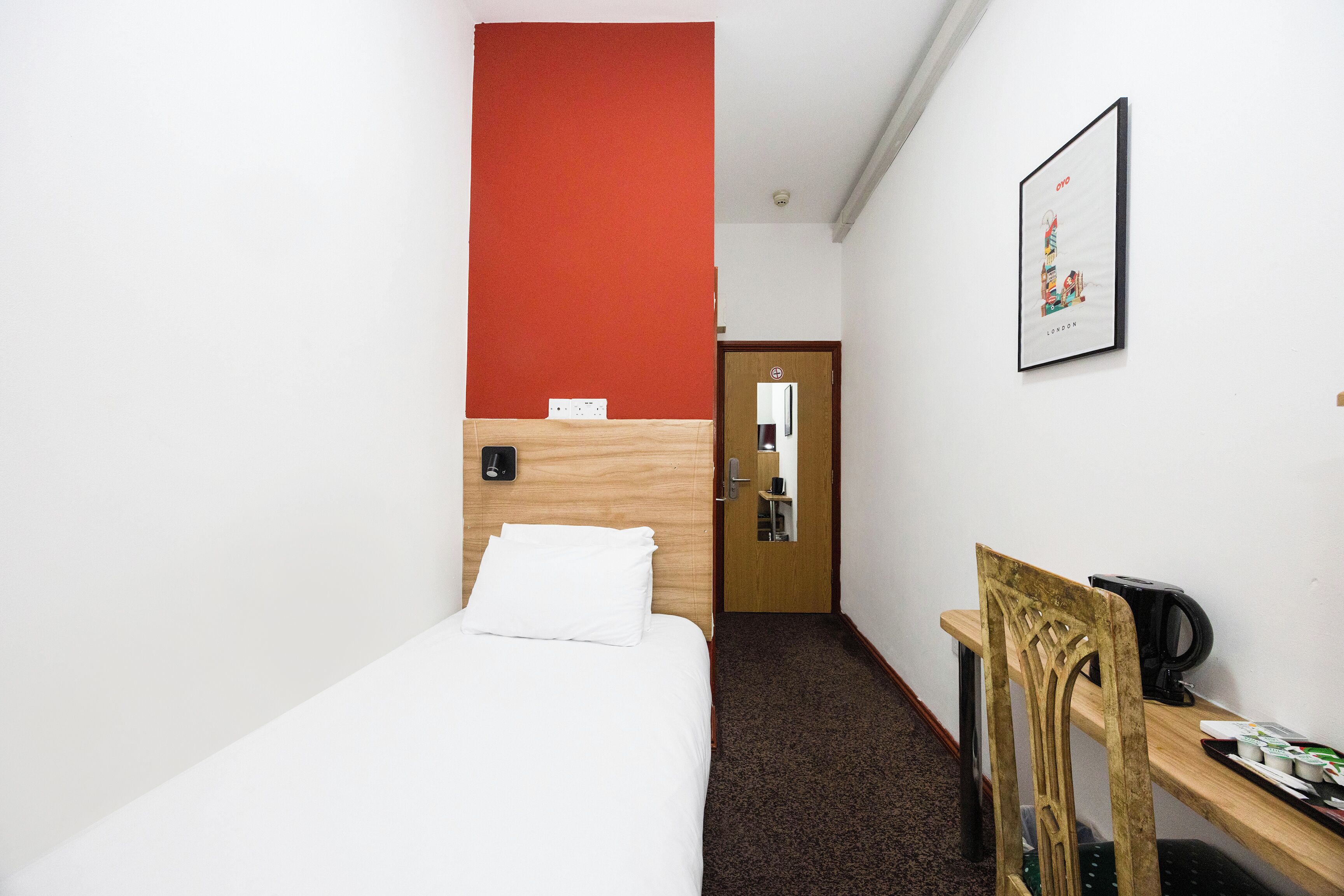 Een hotelkamer met een eenpersoonsbed, een houten bureau, een stoel, een lijst met een afbeelding aan de muur, en een deur naar een badkamer.