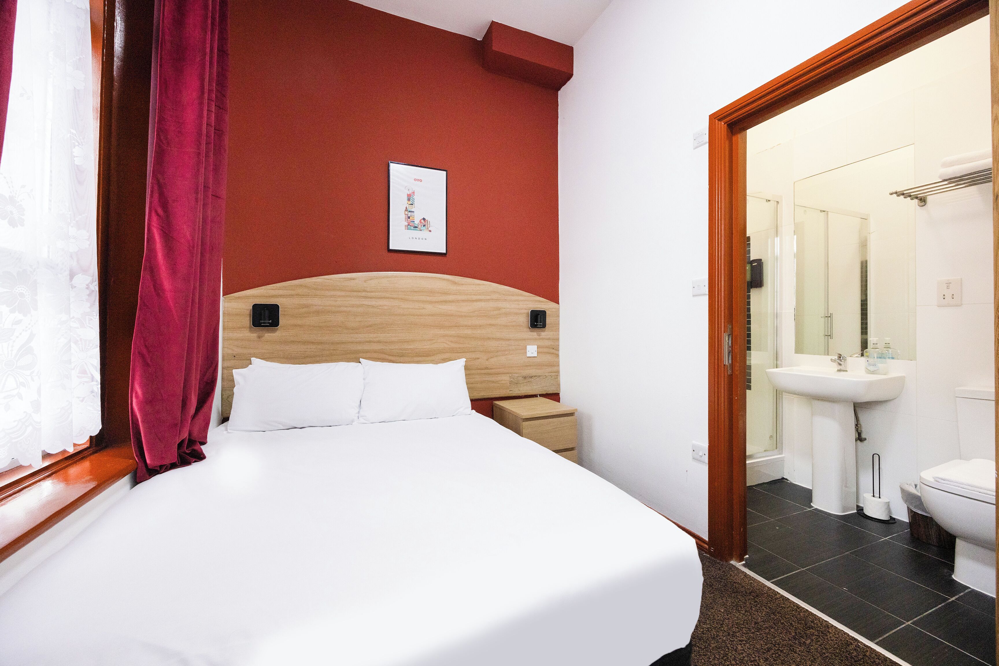 Habitación de hotel con cama de madera, ropa de cama blanca, pared de acento roja, un cuadro enmarcado, una mesita de noche, un pequeño cajón, un baño con lavamanos e inodoro, y un espejo.