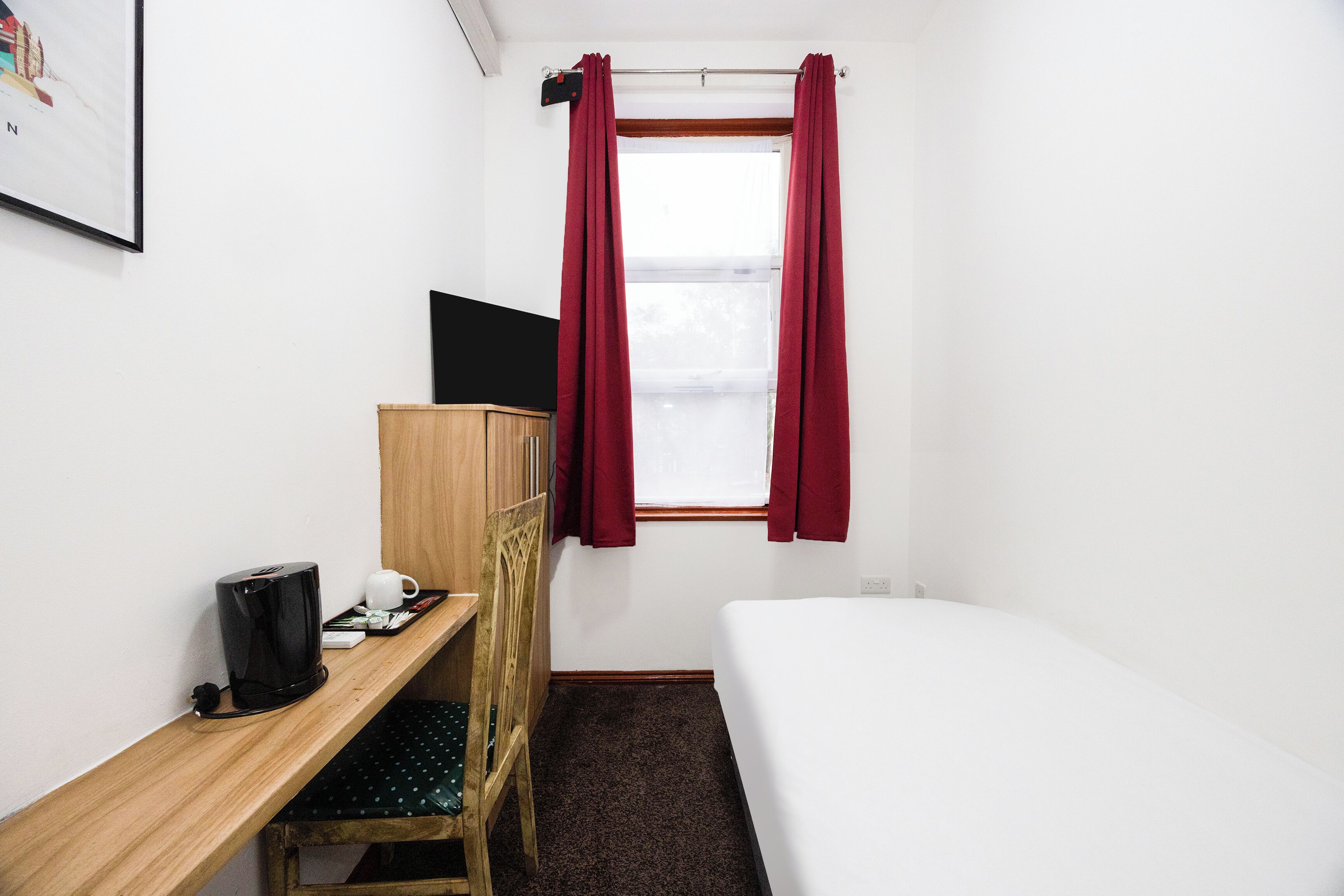 Ett litet hotellrum med en enkel säng, ett skrivbord i trä med en stol, en svart kaffebryggare och en tv.