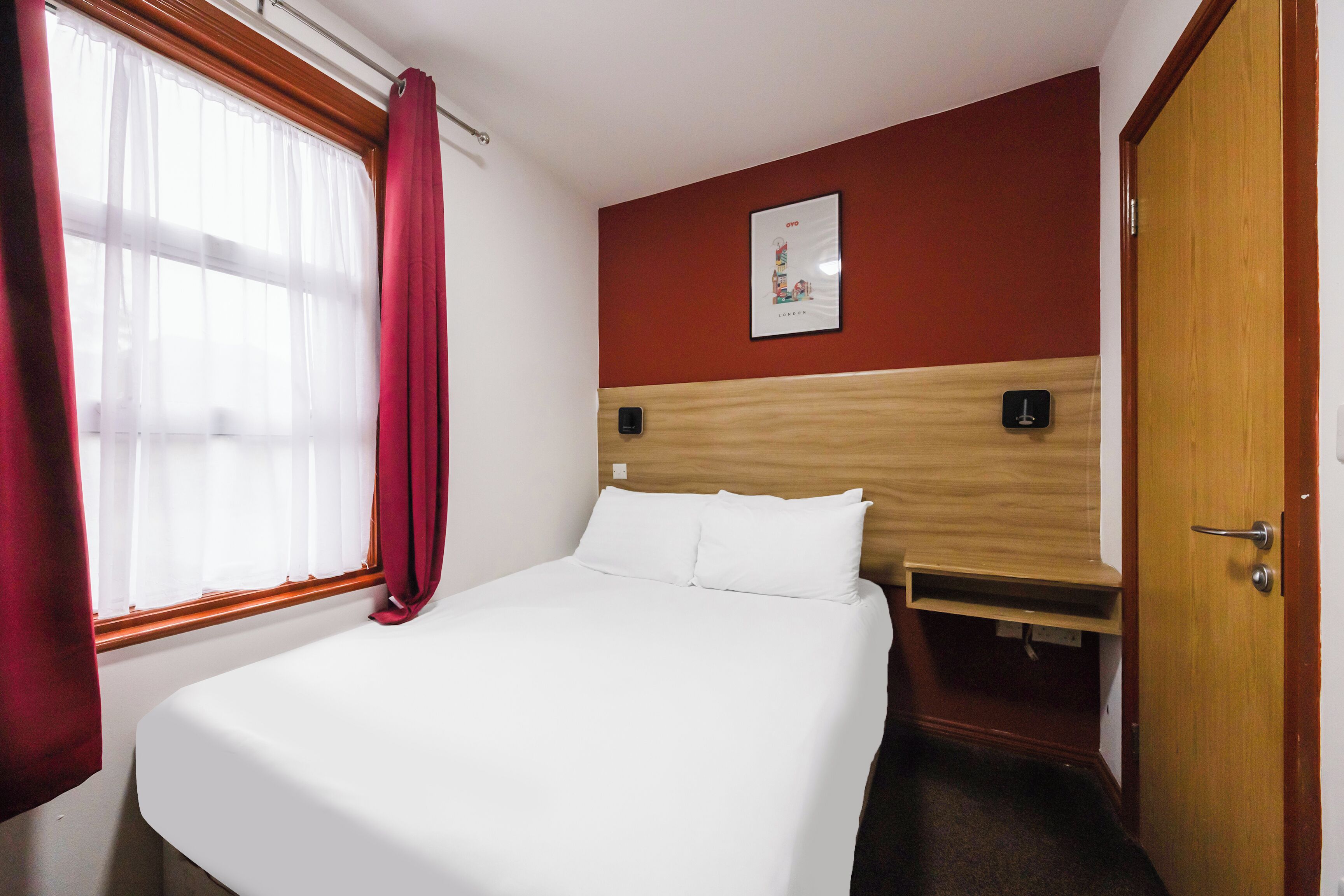 A hotel room with a bed, a wooden headboard, a window with curtains, a door, and a wall-mounted light switch.