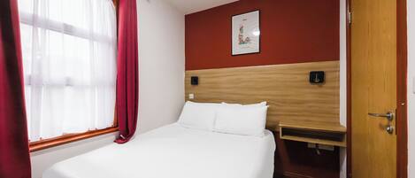 A hotel room with a bed, a wooden headboard, a window with curtains, a door, and a wall-mounted light switch.