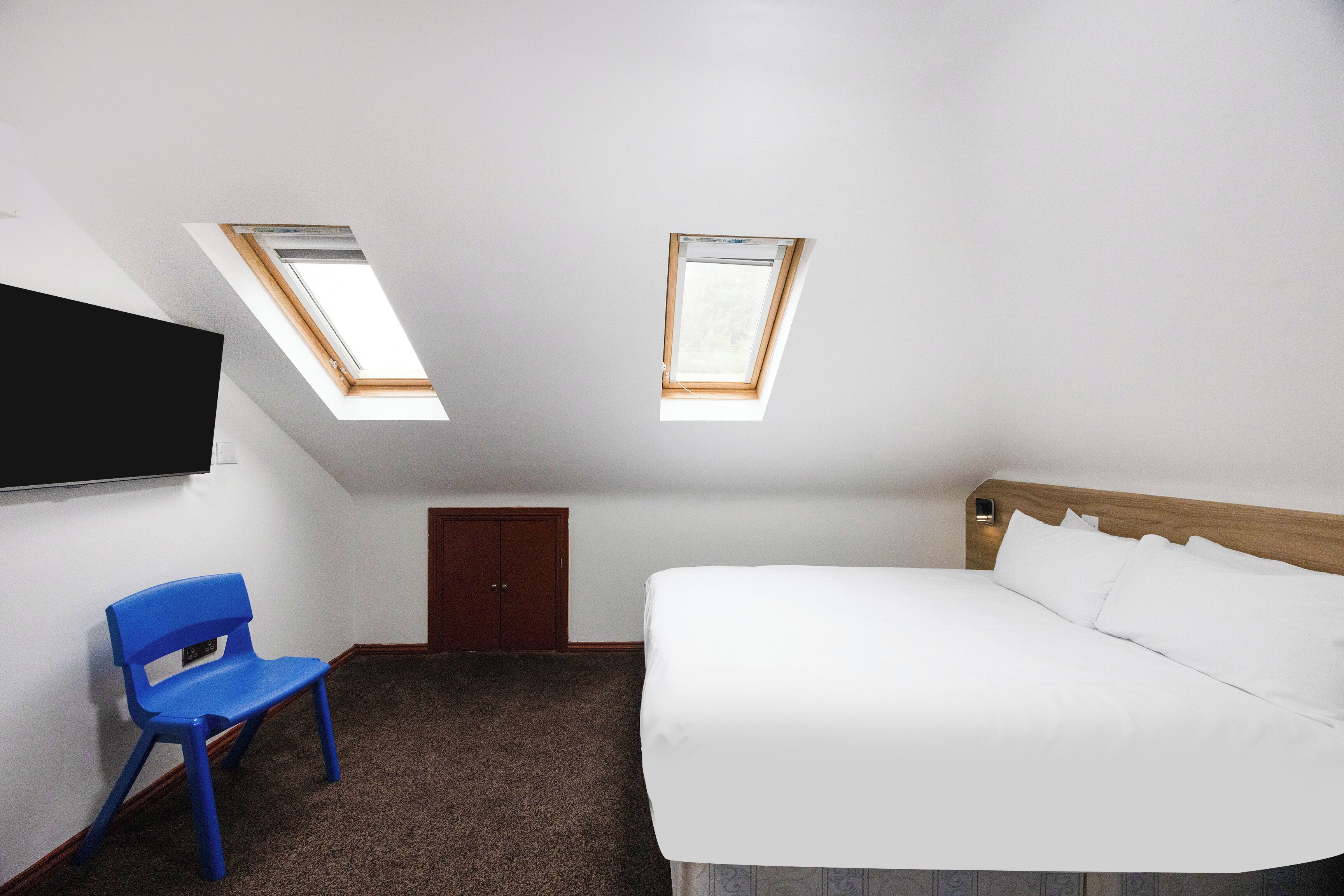 A bedroom with a bed, a blue chair, a wooden headboard, a TV mounted on the wall, and two skylights.