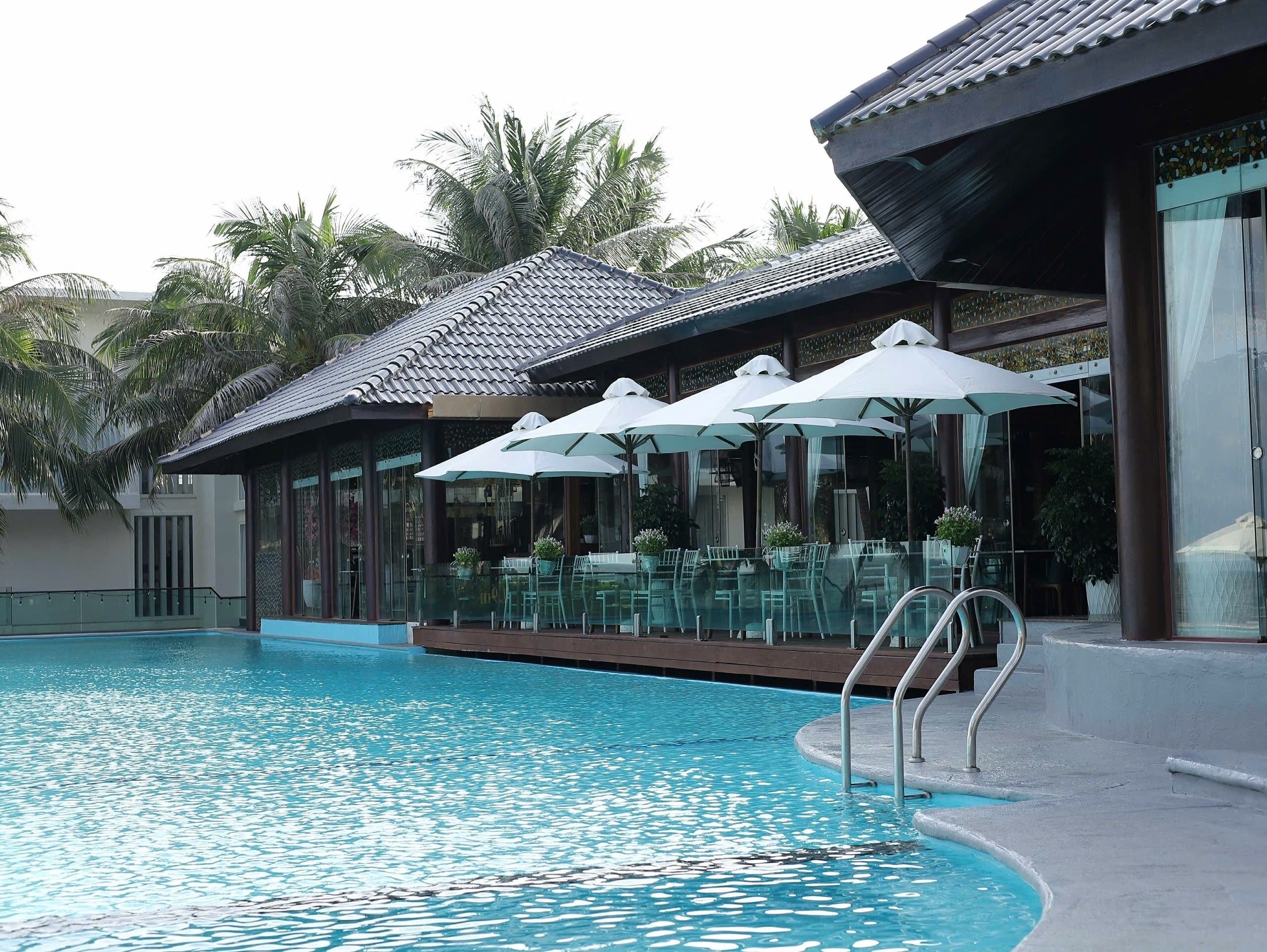 A swimming pool with clear blue water, surrounded by lounge chairs and umbrellas, and a building with a tiled roof and large windows.