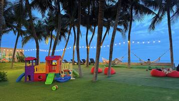 A children's play area with a slide and a small train, surrounded by palm trees and string lights.