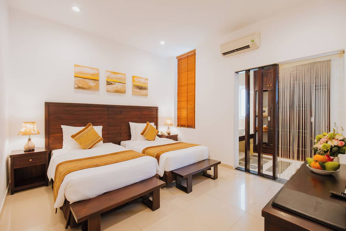 A hotel room with two beds, a wooden headboard, a bench, a desk with a vase of flowers, and a sliding door leading to a balcony.