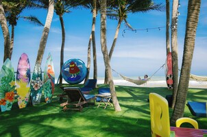 Zona de playa con sillas de descanso, una hamaca y tablas de surf, con palmeras y el océano de fondo.