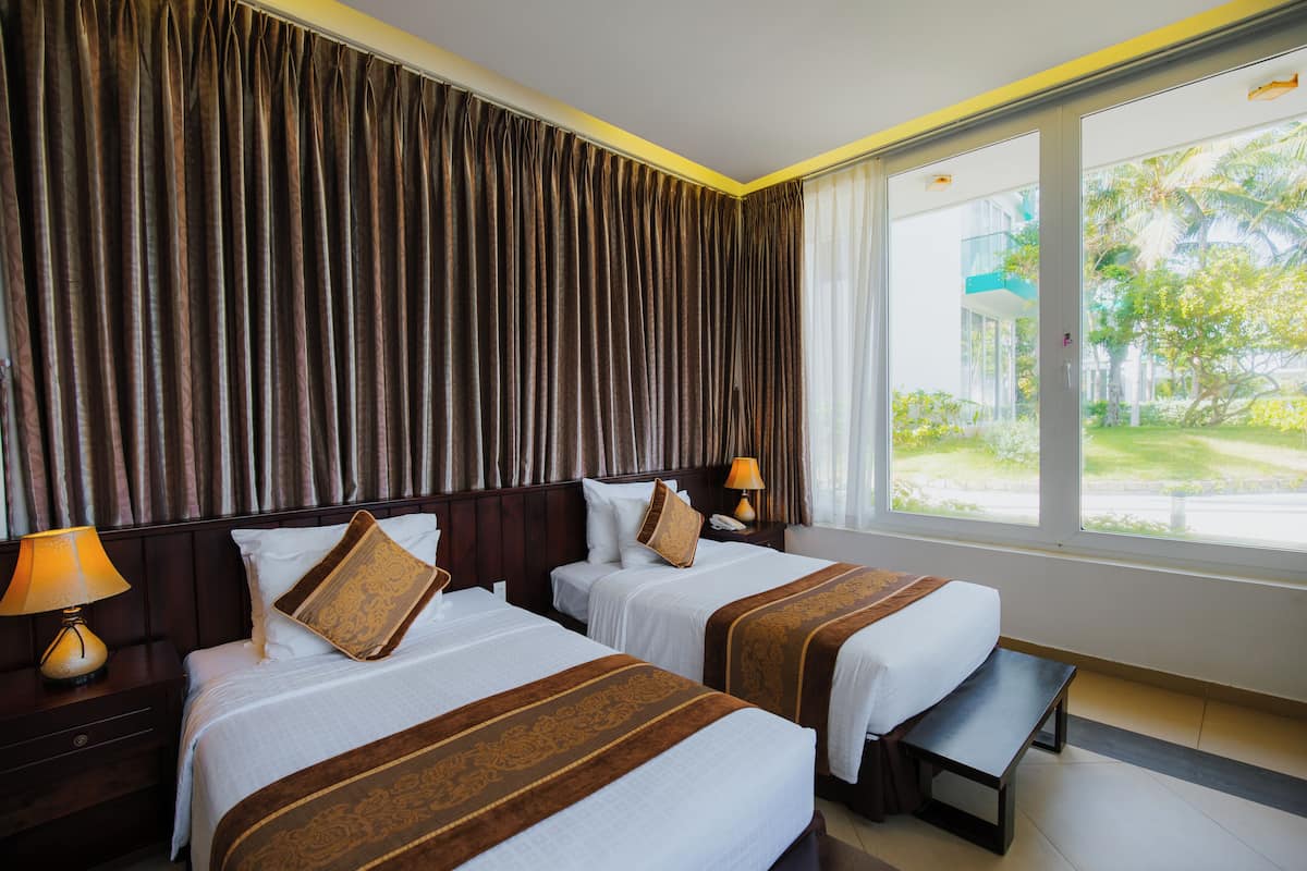 A hotel room with two beds, a bench, a nightstand with a lamp, and a window with a view of greenery.