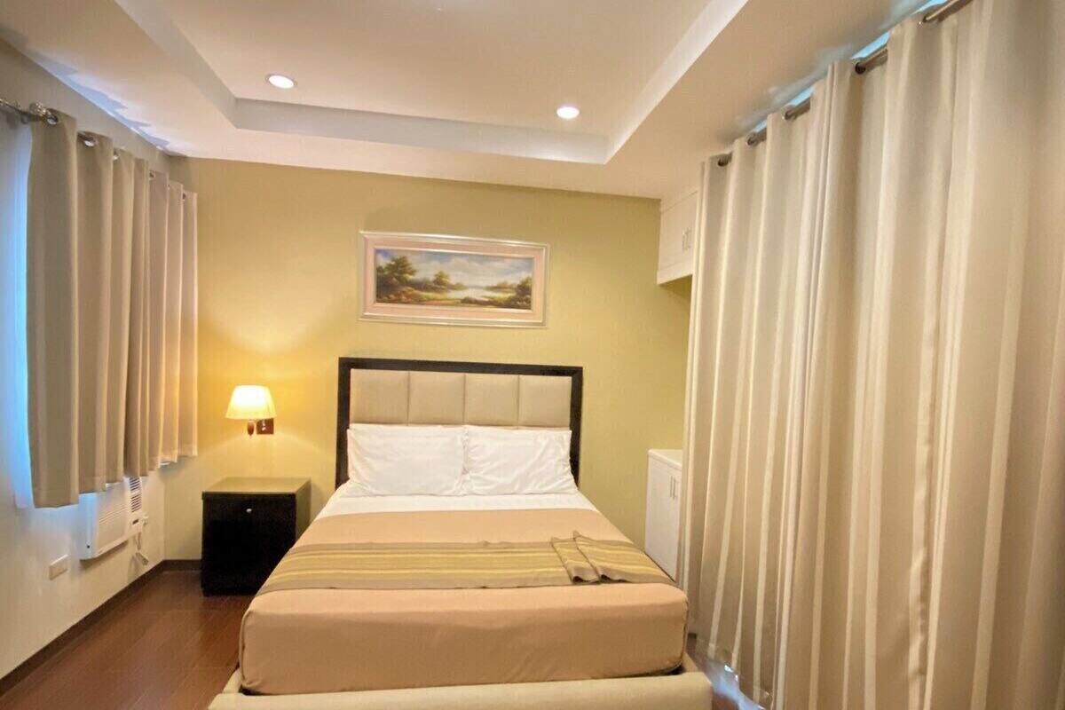 Una habitación de hotel con una cama, una mesita de noche, una lámpara, un cuadro en la pared y cortinas en las ventanas.