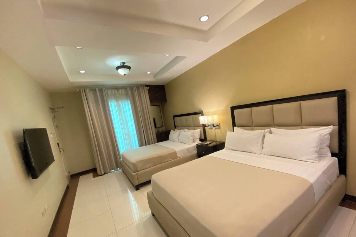 Habitación de hotel con una cama grande, una cama más pequeña, un escritorio con lámpara, ventilador de techo y televisor montado en la pared.