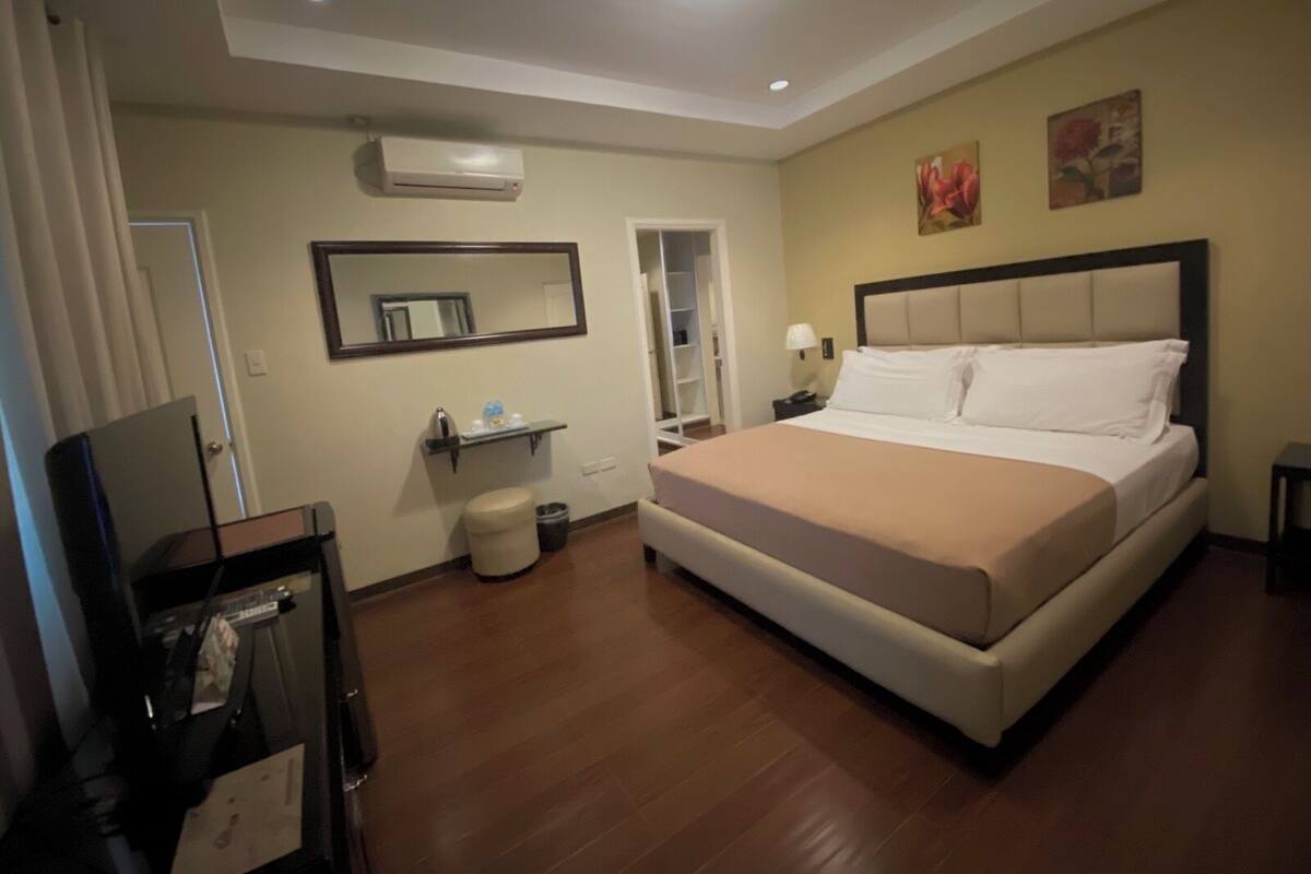 Habitación de hotel con cama, televisión, espejo, repisa con artículos de aseo, cubo de basura y un cuadro en la pared.