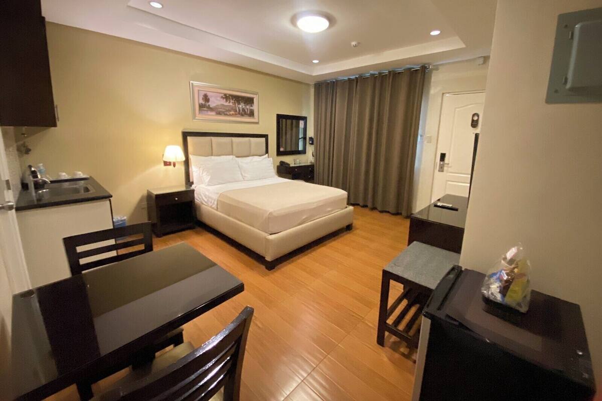 Habitación de hotel con cama, escritorio, silla, mesita pequeña, mesita de noche, lámpara, un cuadro en la pared, ventana con cortinas y puerta.