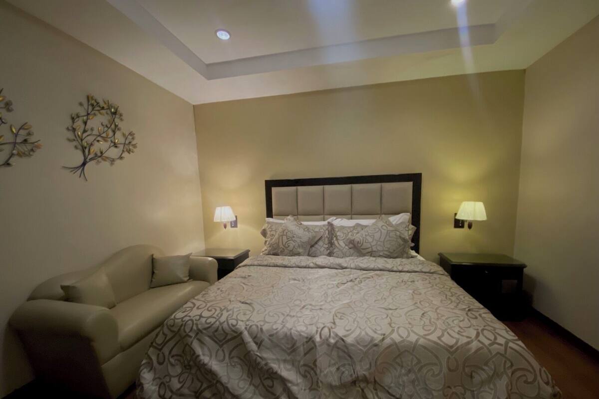Un dormitorio con cama, dos mesitas de noche, un sofá y decoración en la pared.