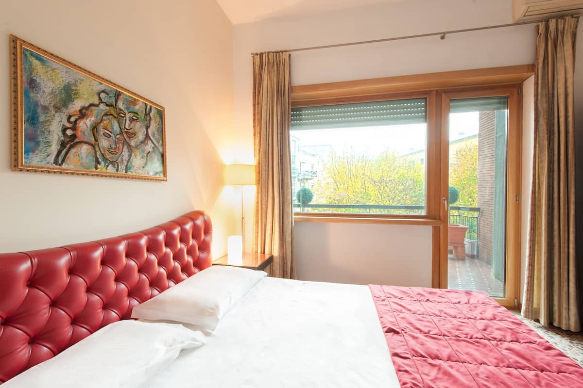 Ein Hotelzimmer mit einem roten, gepolsterten Kopfteil, einem großen Fenster mit Blick auf Bäume und einem Gemälde an der Wand.