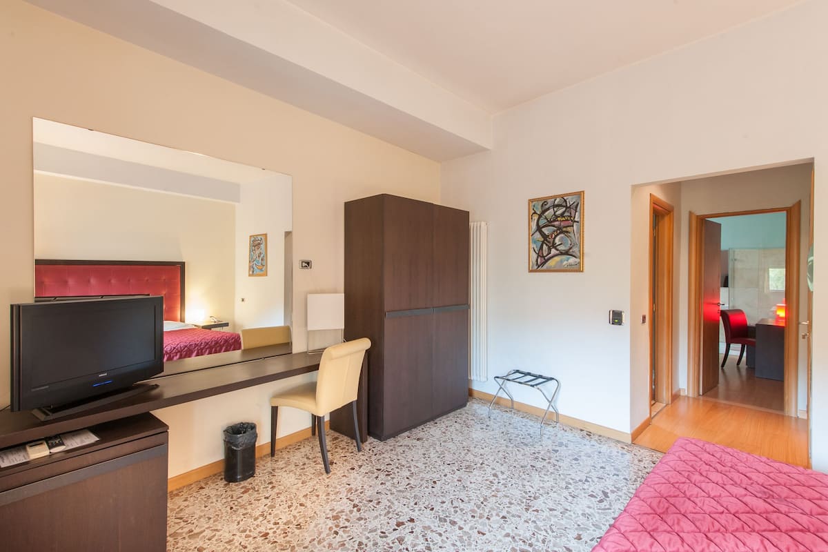 Ein Hotelzimmer mit einem Bett, einem Schreibtisch mit Stuhl, einem Fernseher, einem Kleiderschrank und einem kleinen Tisch mit Sitzgelegenheit.