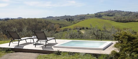 A wooden deck with a rectangular pool and two chairs facing a scenic view of rolling hills and a distant town.
