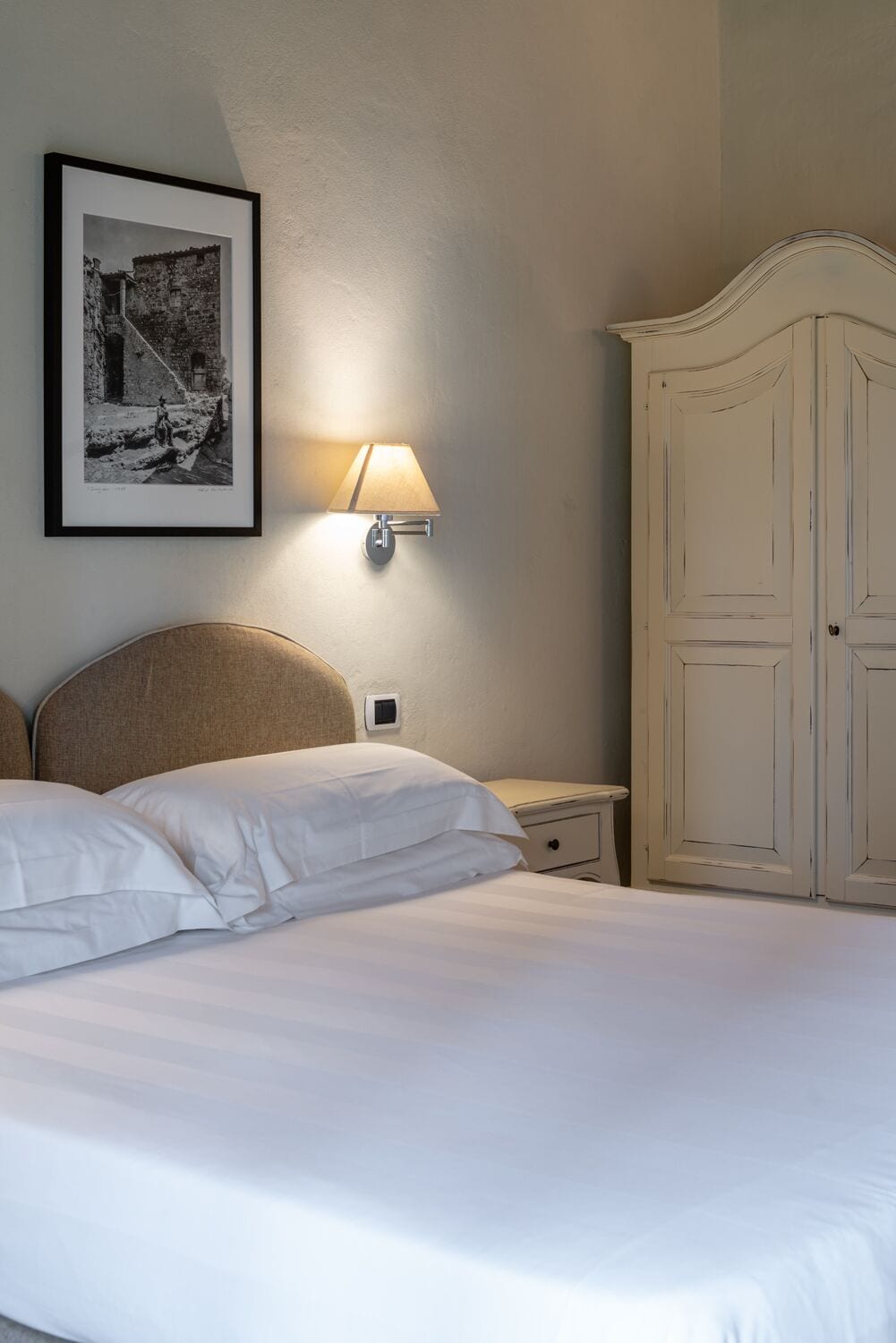 Una cama bien tendida con sábanas blancas, una lámpara de pared y una fotografía histórica enmarcada encima.