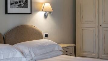 Una cama bien tendida con sábanas blancas, una lámpara de pared y una fotografía histórica enmarcada encima.
