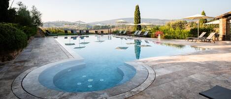 Una piscina con un jacuzzi en el centro, rodeada de sillones y sombrillas.
