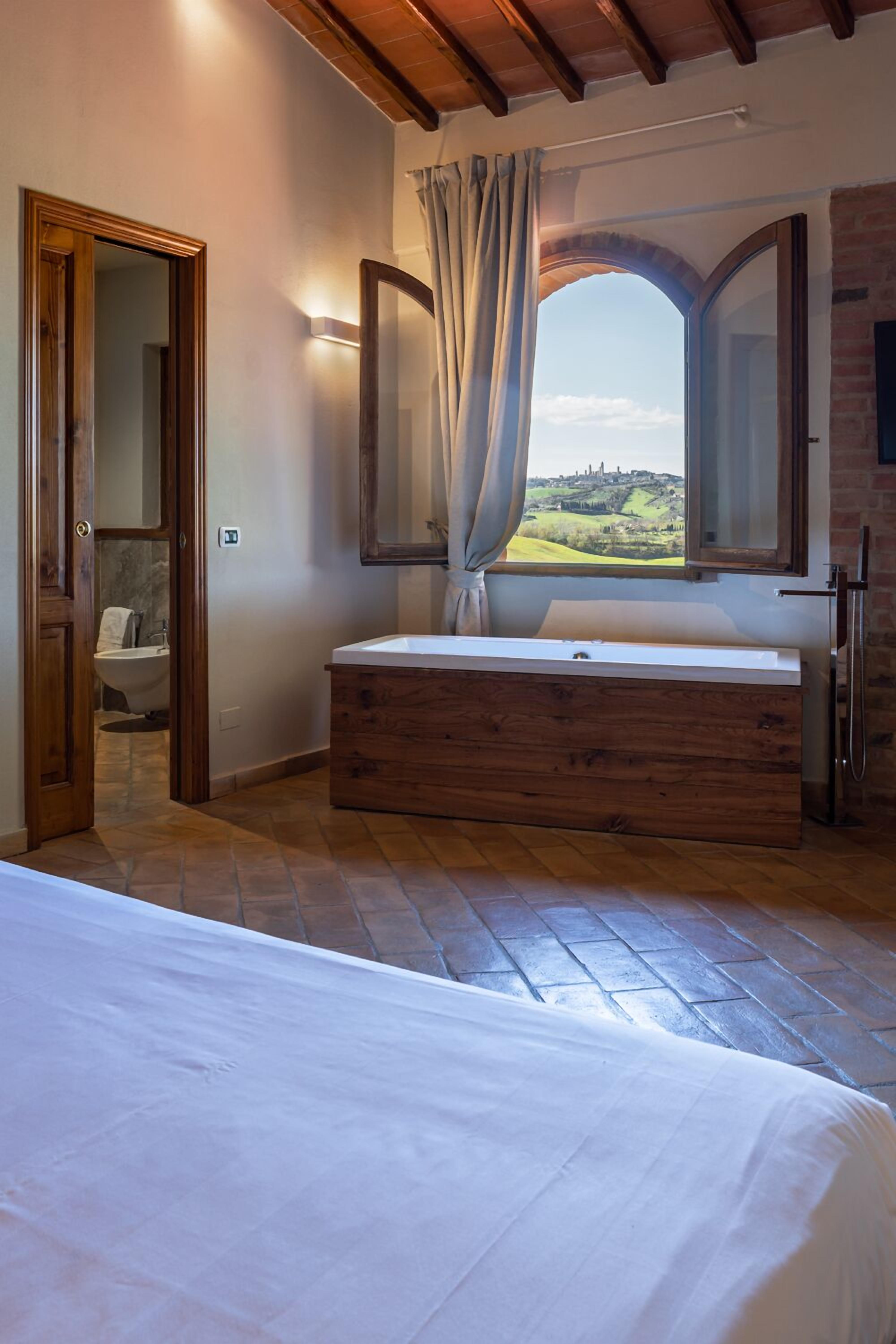 Un dormitorio con cama de madera, un ventanal con vistas, tocador de madera y un baño visible a través de una puerta abierta.