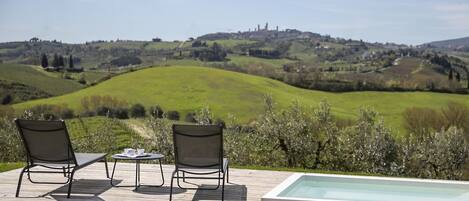 Zona de piscina exterior con dos sillones de descanso y una mesa, con vistas a un paisaje montañoso y un castillo distante.